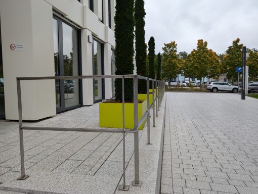 Rampe mit Edelstahlgeländer an einem modernem Gebäude, umgeben von Pflanzen und Parkplatz.