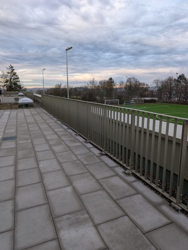 Blick auf einen gepflasterten Gehweg und Sportplatz unter bewölktem Himmel.